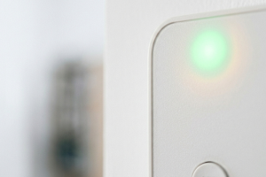 Plano de detalle en primer plano de un panel o teclado de alarma de color blanco con una luz led indicadora encendida en color verde, que muestra que el sistema de seguridad de la vivienda está activo, con un fondo interior desenfocado.