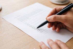Primer plano de una mano sosteniendo un bolígrafo negro sobre un formulario impreso en papel blanco, apuntando a una lista de casillas de verificación para marcar, simulando la solicitud del bono social eléctrico sobre un escritorio.