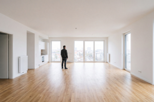 Un hombre de espaldas en el centro de un piso amplio, vacío y luminoso con suelo de madera clara y paredes blancas, mirando hacia los grandes ventanales del fondo que muestran vistas urbanas de fondo, con una cocina americana integrada a la izquierda.