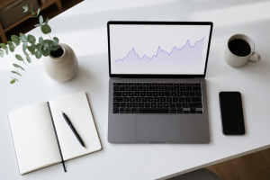 Vista cenital de un portátil abierto que muestra un gráfico lineal ascendente con la evolución del precio de la luz, sobre un escritorio blanco con un cuaderno, un bolígrafo, un teléfono móvil y una taza de café, simulando el análisis de la tarifa regulada PVPC de Curenergía.