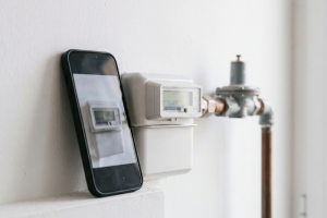 Un teléfono móvil con funda negra apoyado junto a un contador de gas digital e instalado en una pared blanca, mostrando en su pantalla la fotografía del propio medidor para enviar la lectura a Nedgia.