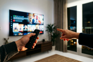 Primer plano de las manos de dos personas en un salón por la noche; una sostiene un mando a distancia negro apuntando hacia una televisión inteligente grande colgada en la pared que muestra un catálogo de películas en streaming, mientras la otra persona señala la pantalla.