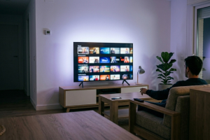 Hombre de espaldas sentado en un sillón gris dentro de un salón iluminado de noche, interactuando con el mando a distancia hacia una televisión inteligente grande que muestra una interfaz de catálogo de películas y series en streaming.