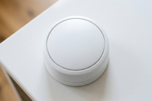 Primer plano cenital de un dispositivo detector o sensor de movimiento inalámbrico de color blanco y forma circular, colocado sobre la esquina de un mueble o superficie clara.