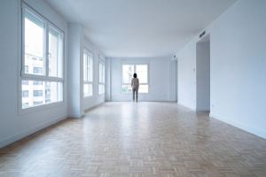 Una persona de espaldas al fondo de una gran estancia completamente vacía y pintada de blanco, con suelo de parqué claro y múltiples ventanales laterales que iluminan el piso recién entregado.