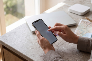 Primer plano en ángulo de las manos de un usuario interactuando con la pantalla táctil de un teléfono móvil inteligente junto a una mesa de mármol claro, donde hay un ordenador portátil cerrado, un router portátil cuadrado blanco y un mapa de carreteras impreso.