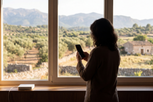 Silueta de espaldas de una mujer sosteniendo un teléfono móvil inteligente frente a un gran ventanal que muestra un paisaje rural montañoso con olivares y una casa de campo, con un pequeño router portátil blanco colocado sobre el marco inferior de la ventana.