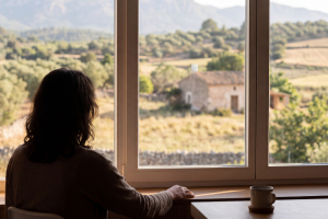 Silueta desde los hombros de una mujer sentada frente a un gran ventanal que mira hacia un paisaje de campo con olivares y una casa rústica al fondo. En el cristal de la ventana hay adherida una pequeña antena receptora blanca con un cable, y en primer plano a la derecha, una taza sobre una mesa de madera.