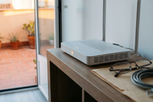 Router wifi de color blanco con antenas sobre un mueble de madera cerca de unas gafas y un cable de red.