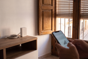 Persona comprobando la señal WiFi en su móvil frente a un repetidor de señal blanco sobre un mueble de madera.
