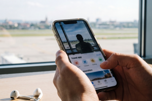 Manos sosteniendo un teléfono móvil con una aplicación de viajes frente al ventanal de un aeropuerto.
