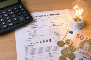 Factura de electricidad sobre una mesa junto a una calculadora, monedas y billetes de euro, y una bombilla encendida.