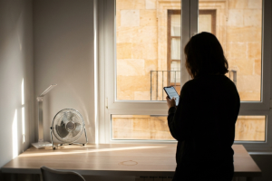Silueta de una mujer de espaldas usando su smartphone junto a un escritorio con ventilador y una ventana iluminada.