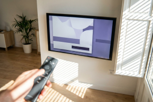 Primera persona de una mano sosteniendo un mando a distancia hacia una televisión Smart TV colgada en la pared del salón.
