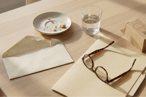 Vista superior de un sobre abierto, gafas con montura de carey sobre un folio, un vaso de agua y un plato con migas en un escritorio de madera.