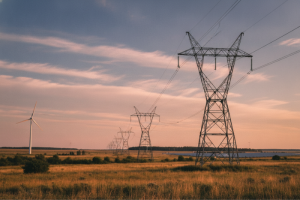 Línea de torres eléctricas de alta tensión cruzando un campo abierto junto a un aerogenerador bajo un cielo con nubes al atardecer.